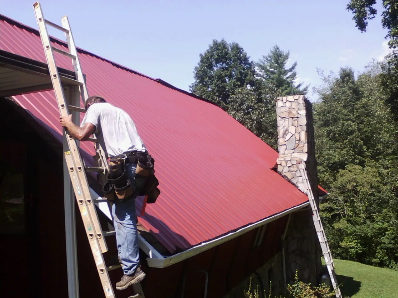 Roofer on a ladder beside a red metal roof and stone chimney — Soffit & Fascia Repair by Roofing Brothers in Kyburz